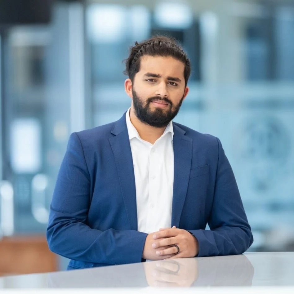 A man wearing a blue suit and white shirt, with dark hair and a beard, standing in a modern office environment with blurred glass windows in the background.