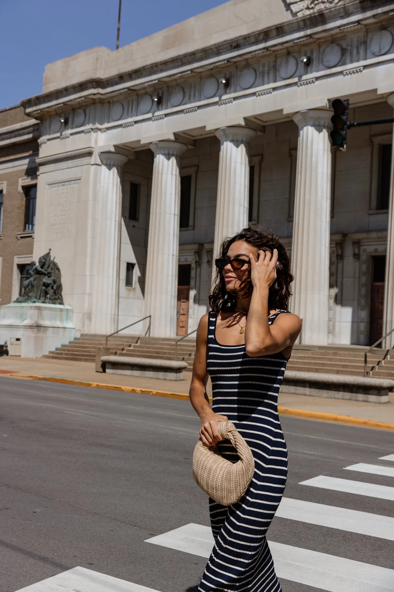 A woman in a black and white striped dress and sunglasses walking across a crosswalk outside a historic building with large columns, a statue, and an ornate facade.