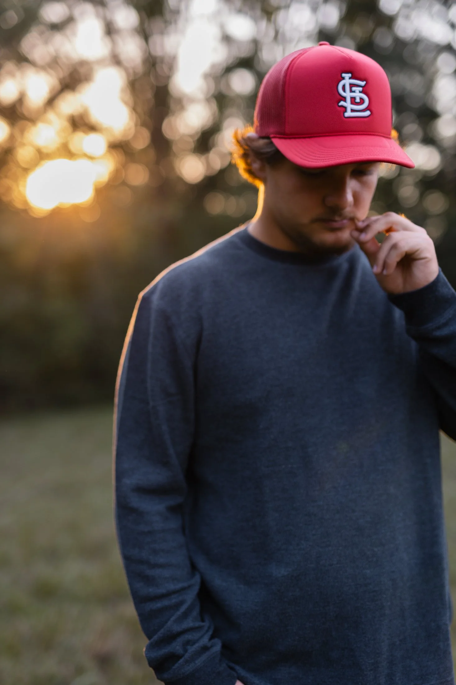 A young man wearing a red baseball cap with a white logo, and a dark long-sleeve shirt, standing outdoors during sunset with a blurred background of trees and warm light.