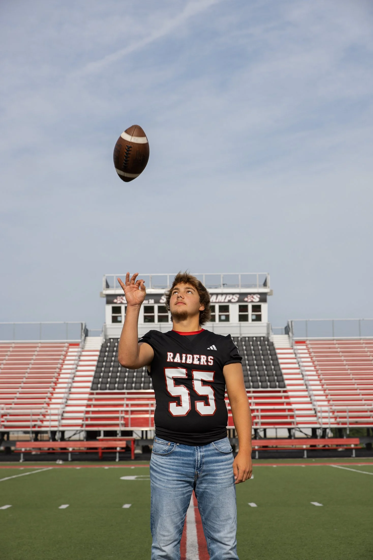 A young male football player in a black and red Raider jersey is standing on a football field and tossing a football into the air, with a stadium in the background.