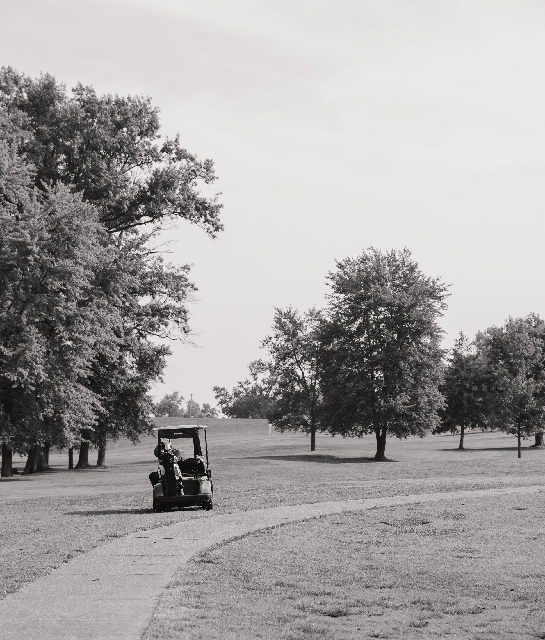 Black and white photo of a golf course with a golf cart and golf bags, surrounded by trees and open grass area.