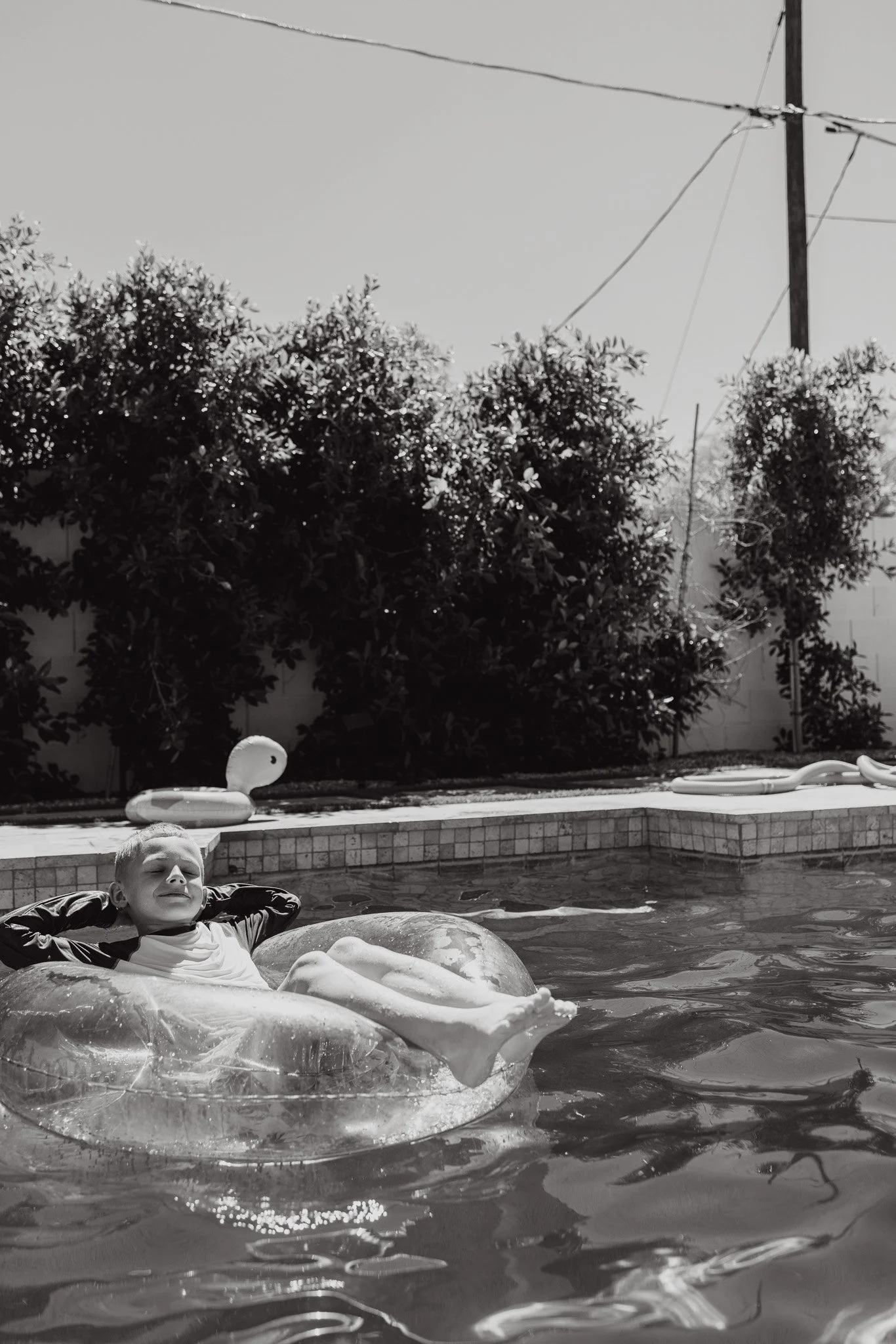 A boy relaxing in a pool on an inflatable float, smiling with eyes closed, with large bushes, a pool deck, and a tall utility pole with wires in the background.