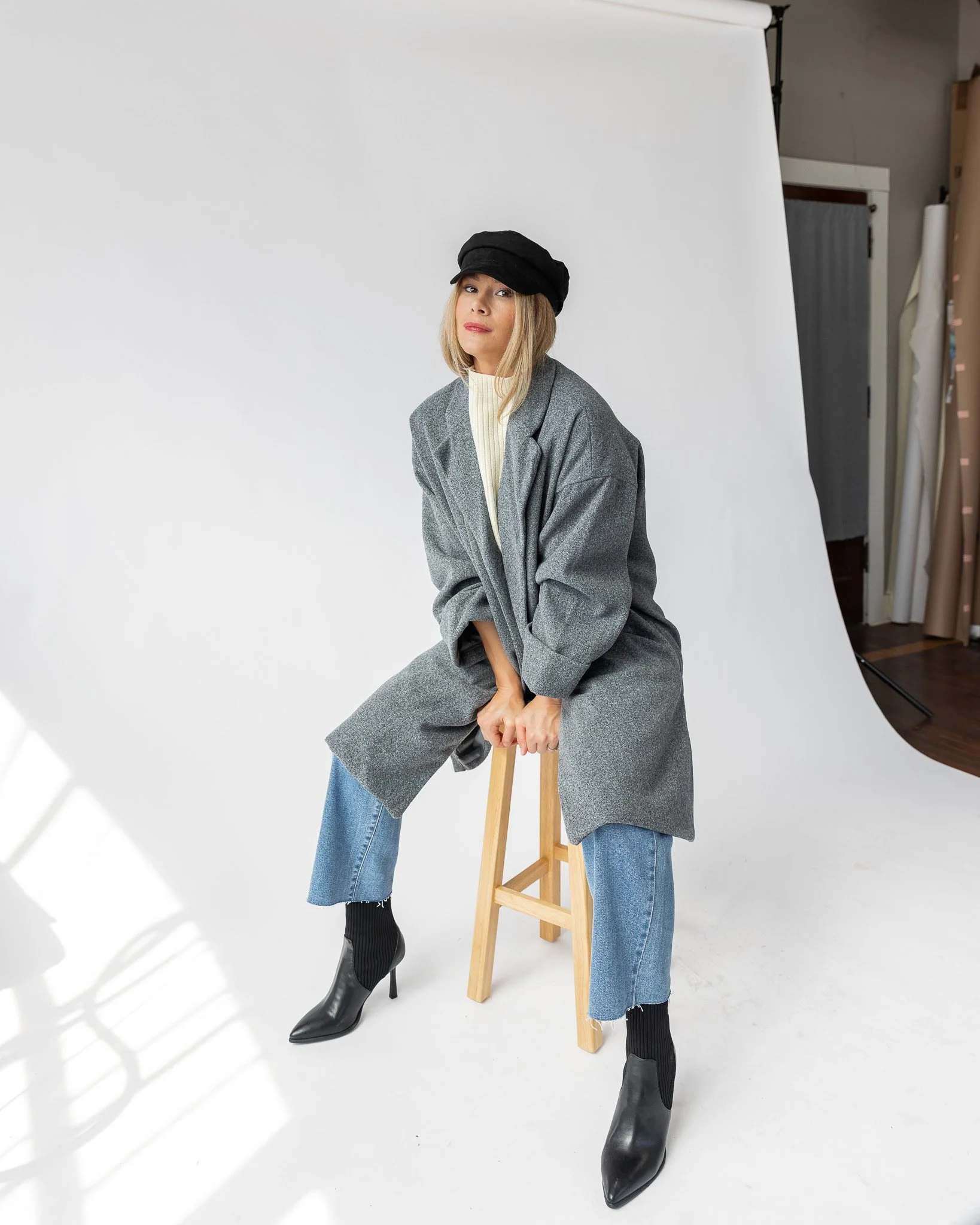 A woman with blonde hair wearing a black cap, grey blazer, light-colored sweater, and blue jeans. She is sitting on a wooden stool with one leg bent and the other leg extended, wearing black ankle boots with high heels. She is in a photo studio with a white background and photography equipment visible.