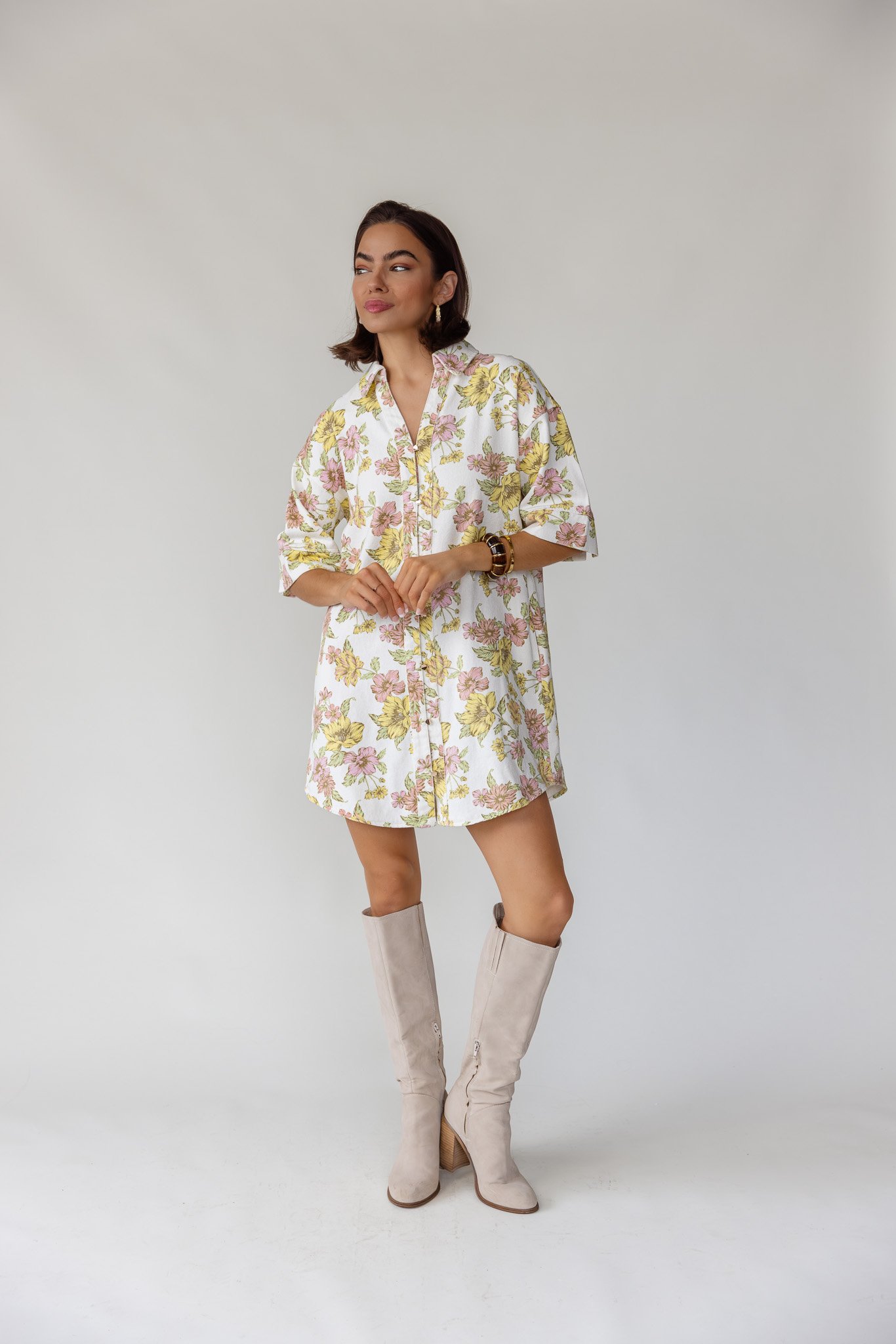 A woman wearing a floral dress, tall beige boots, and a watch, standing against a plain light gray background.