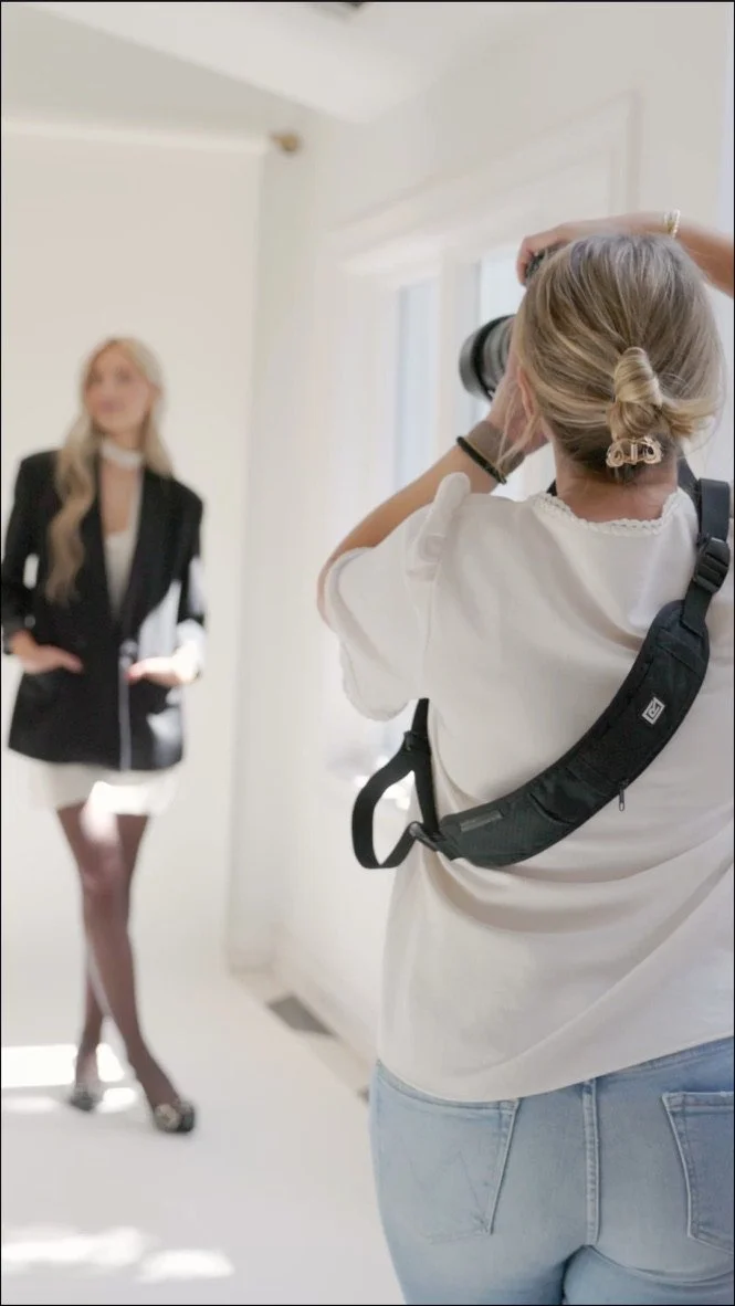 A woman with blonde hair styled in a bun is taking a photograph of another woman in front of a window. The photographer is wearing a white shirt, light blue jeans, and a black camera strap. The woman being photographed is dressed in a black blazer, white skirt, black stockings, and heels, posing with her hands on her hips.
