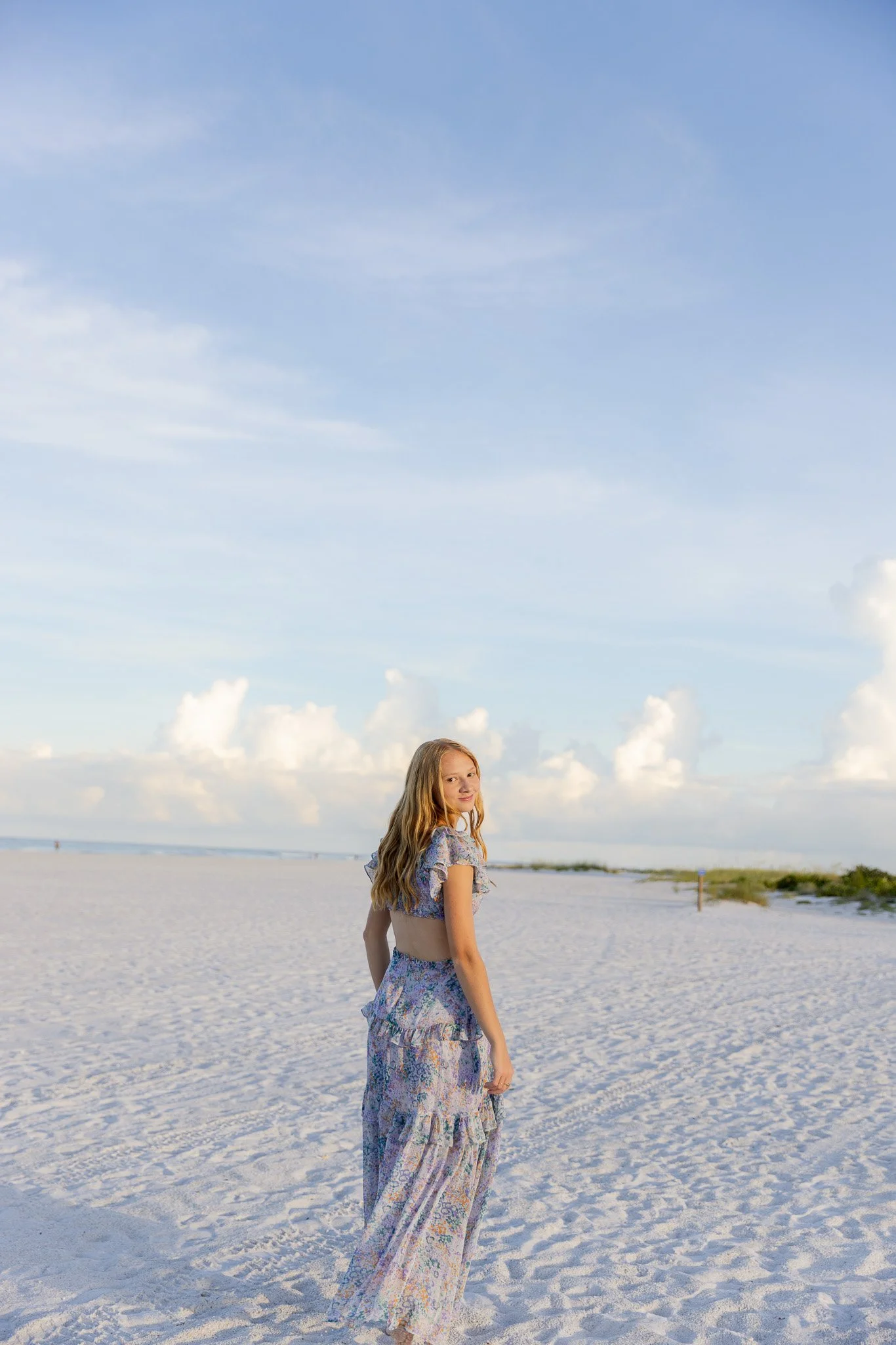 A young woman with long wavy hair standing on a sandy beach, wearing a floral dress, with the ocean and sky with some clouds in the background.