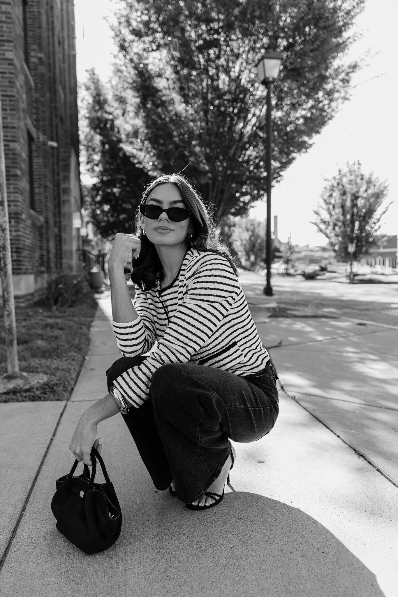 Black and white photo of a woman wearing sunglasses, striped shirt, and sandals, squatting on a sidewalk with a backpack and purse near her feet, with trees, buildings, and street lamps in the background.
