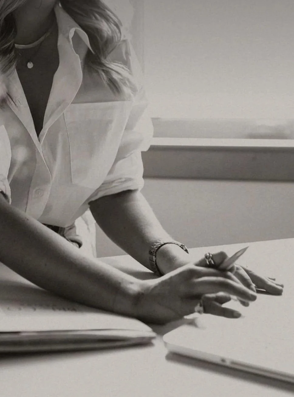 A woman wearing a button-up shirt is sitting at a table, holding and looking at a smartphone with her left hand. There are notebooks or papers on the table.