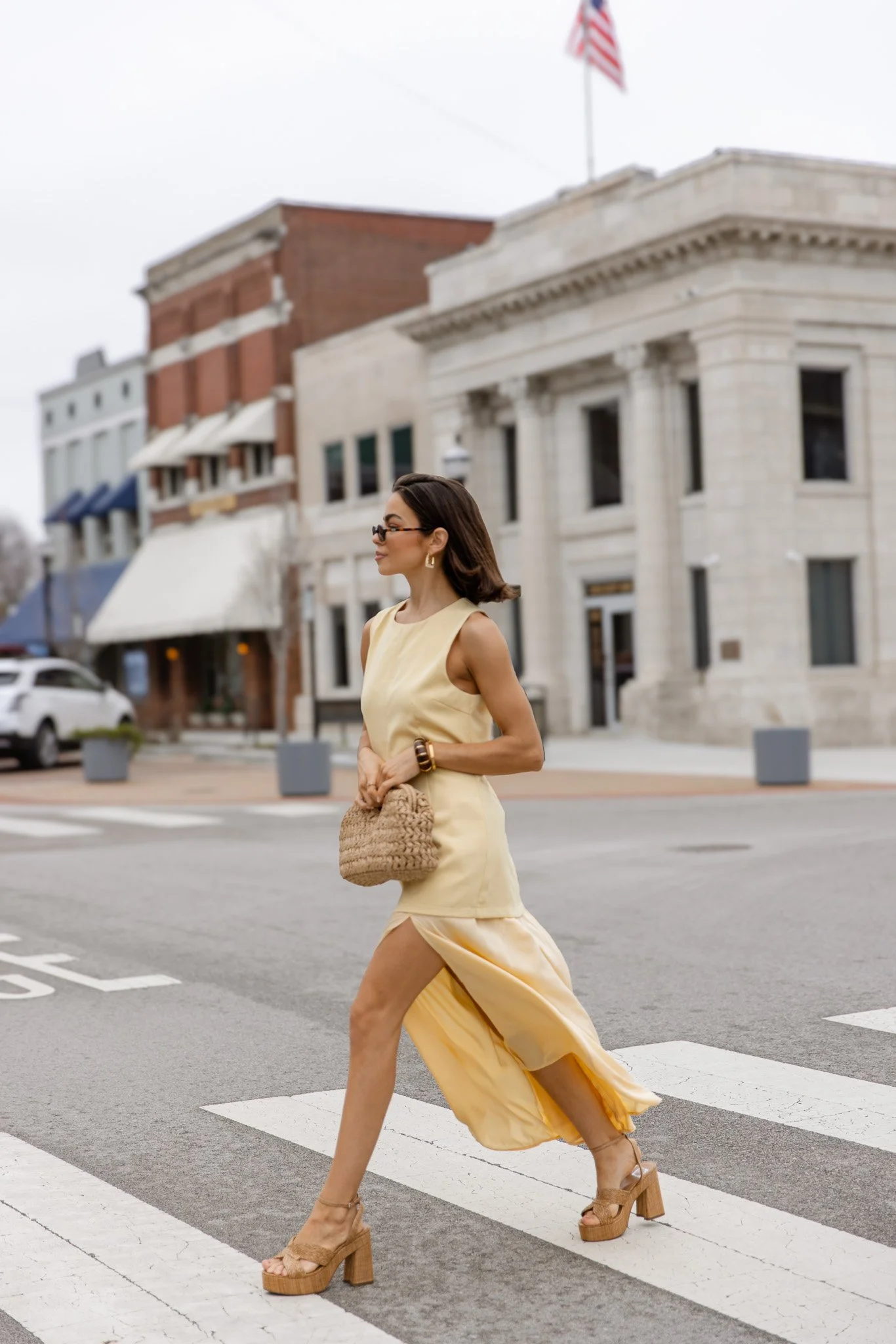 A woman in a yellow dress and platform heels crossing a street in front of historic buildings with an American flag on top.
