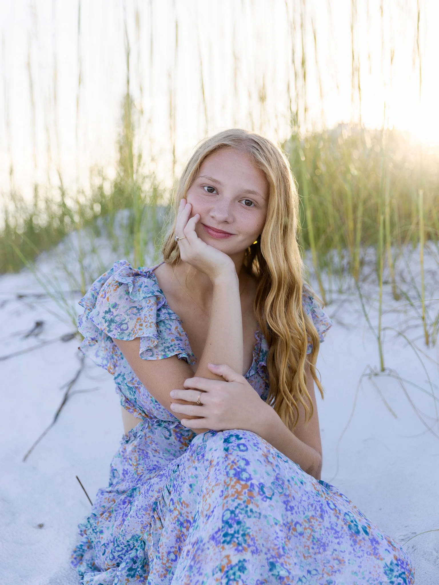 A young woman sitting on white sand at the beach, surrounded by tall green grass, with sunlight glowing behind her, wearing a flowery dress and smiling gently.