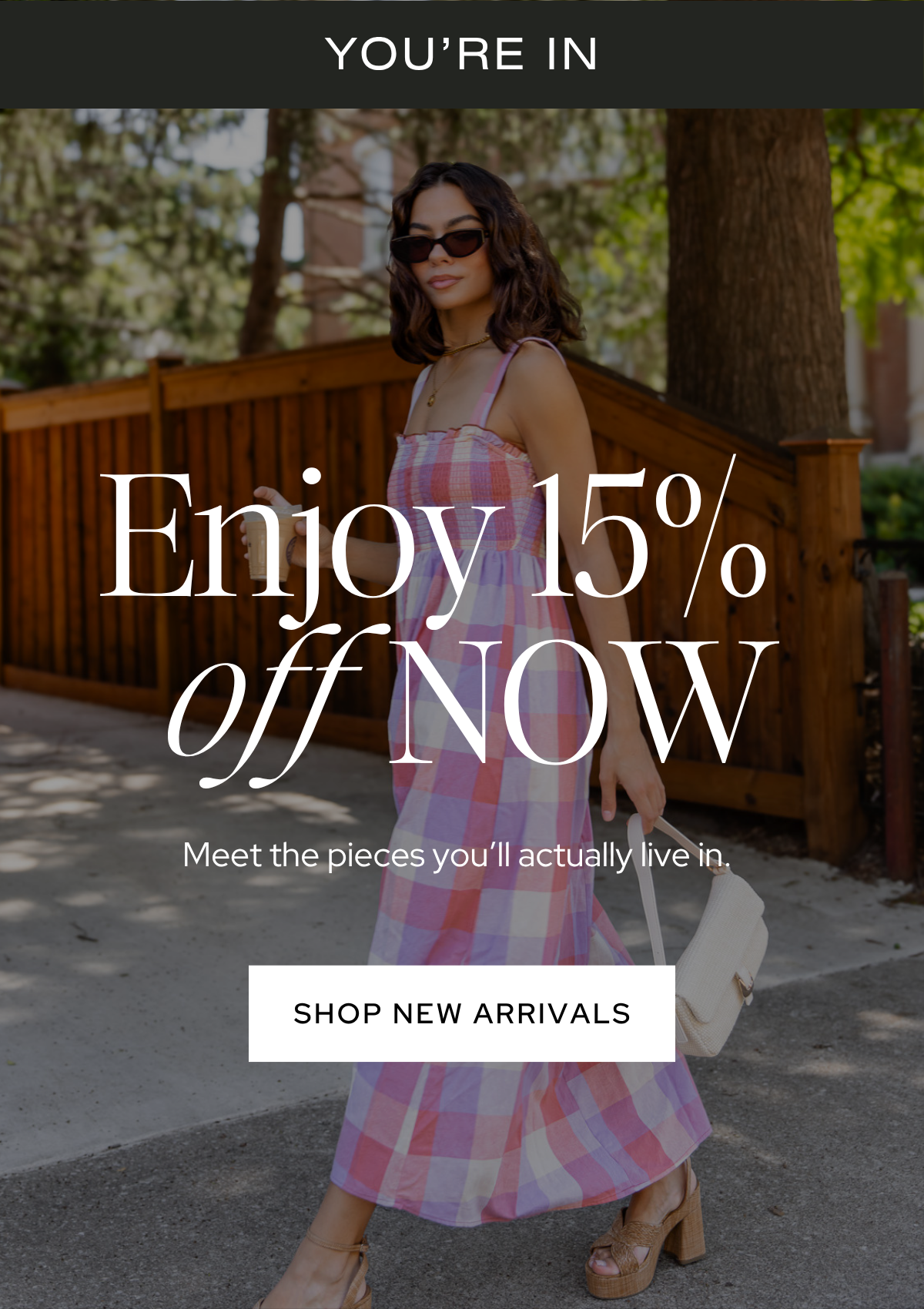 A woman in a pink, purple, and white plaid dress with sunglasses and brown chunky heels standing outdoors near a wooden fence and trees, holding a white bag and a drink, with overlaid text promoting a sale.