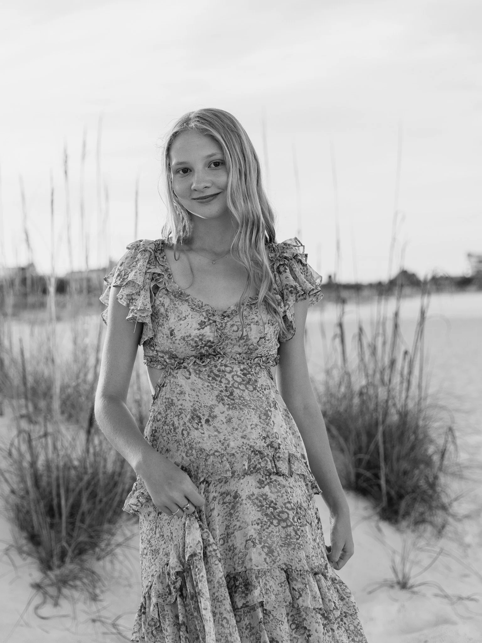 A young woman with long, wavy hair standing outdoors in a natural setting, wearing a patterned dress with ruffled sleeves, with tall grass and a body of water in the background.