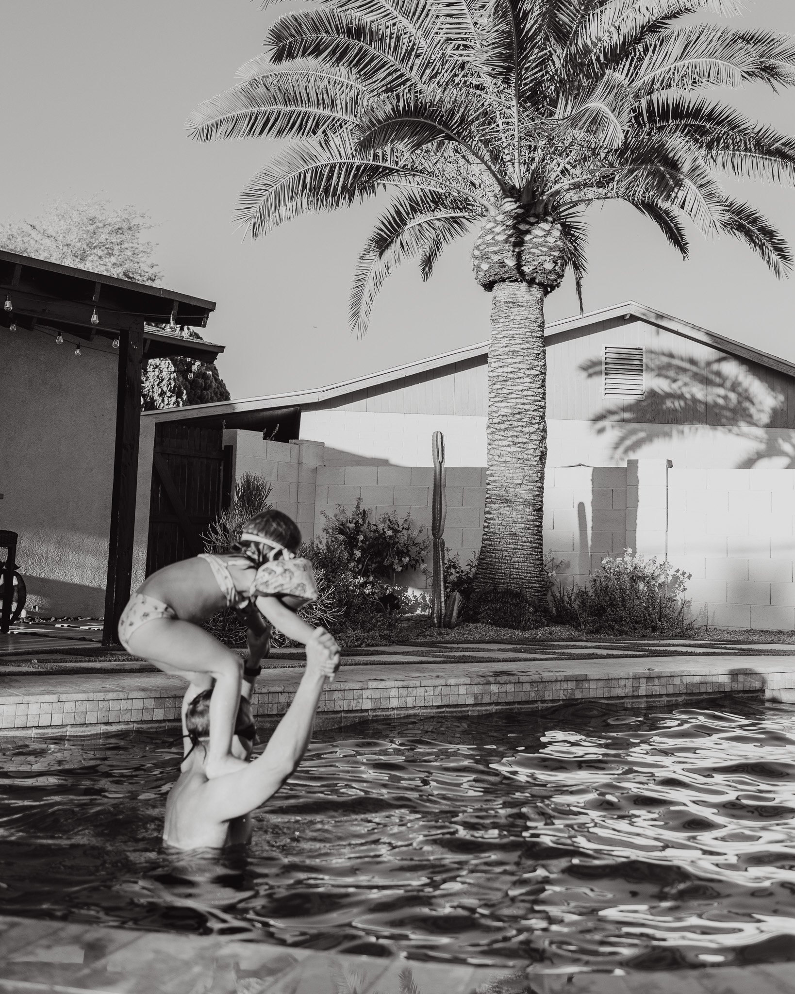 A person diving into a swimming pool with their hands reaching toward the water, a palm tree and house in the background, black and white photograph.