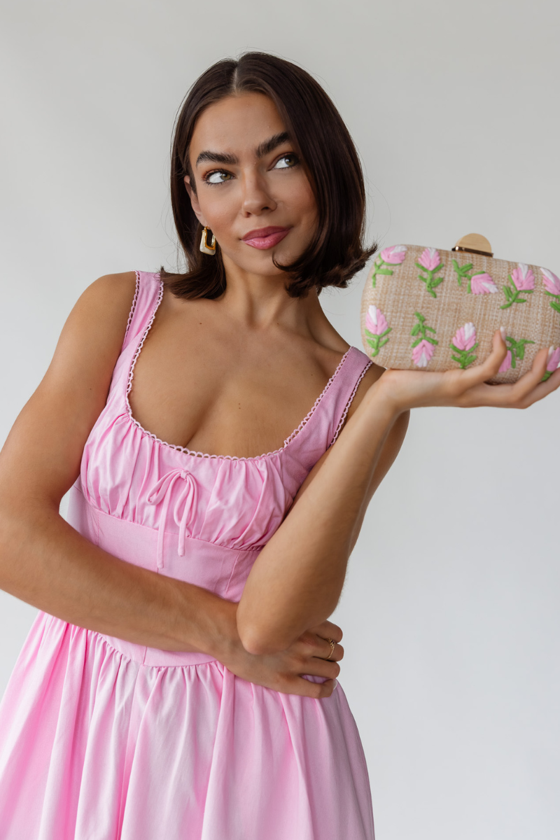 A woman wearing a pink dress holding a floral clutch bag, looking playful and thoughtful.