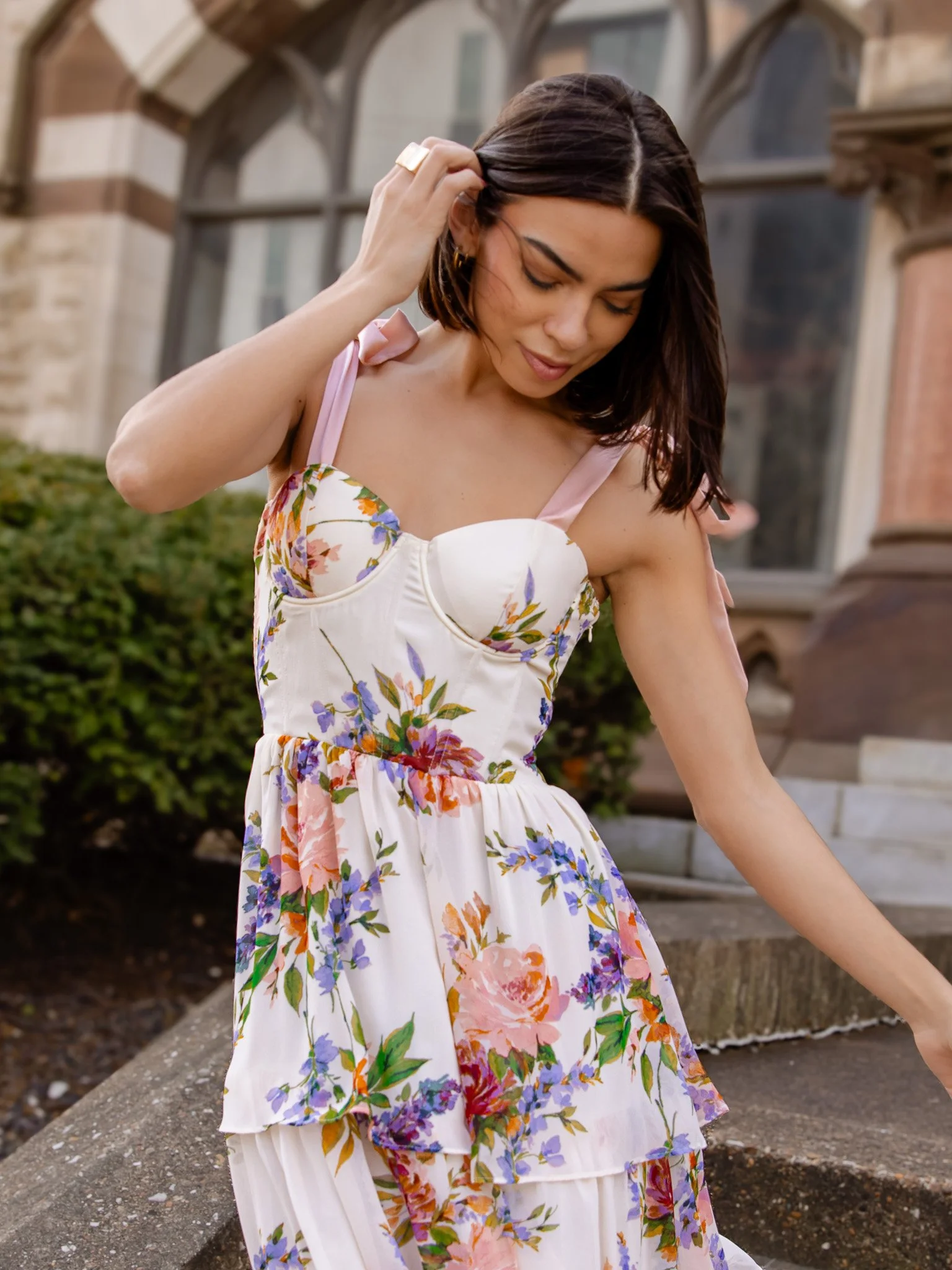 A woman with dark brown hair wearing a floral dress with pink straps, sitting outside near a stone bench and a building with arched windows.