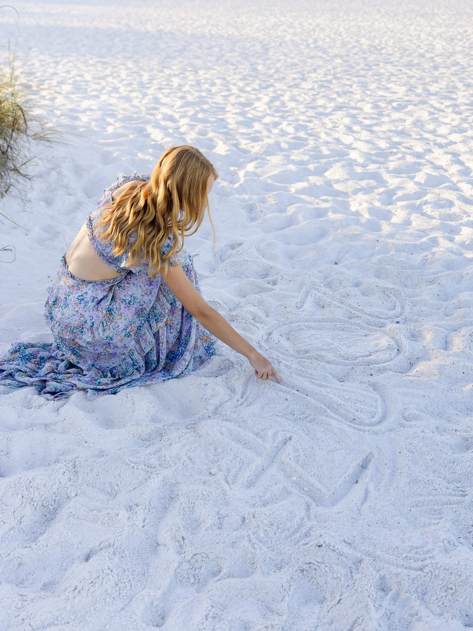 A woman with long, wavy blonde hair wearing a floral dress crouching on the sand at the beach, drawing a heart shape in the sand with her finger. There are footprints in the sand and a patch of grass on the left edge of the image.