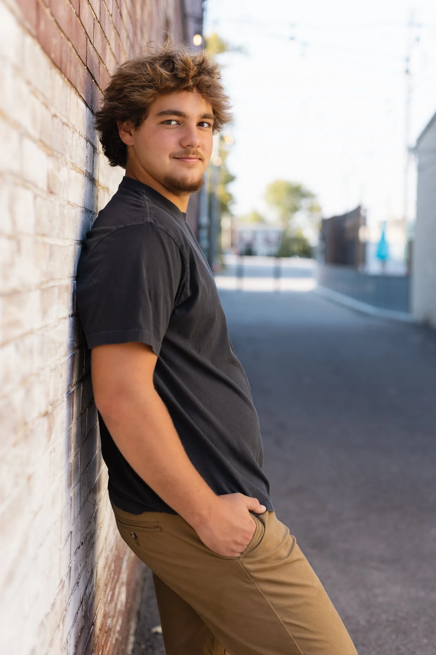A young man with curly brown hair and a goatee leaning against a brick wall in an urban street setting, wearing a black T-shirt and khaki pants, with his hands in his pockets and looking at the camera.