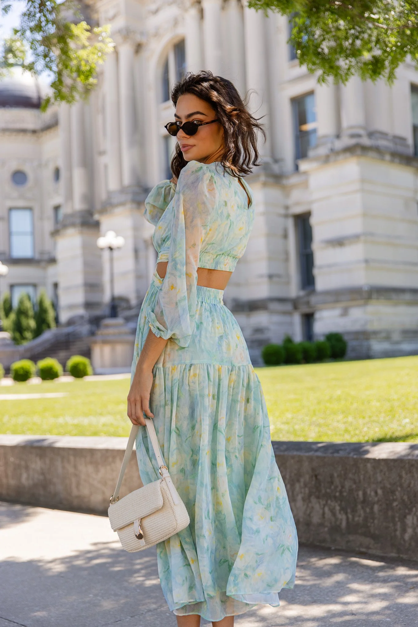 A woman with dark wavy hair wearing sunglasses and a flowy pastel-colored dress with elastic waist, standing outdoors in front of a historic building with large windows, trees, and a well-maintained lawn, holding a white handbag.