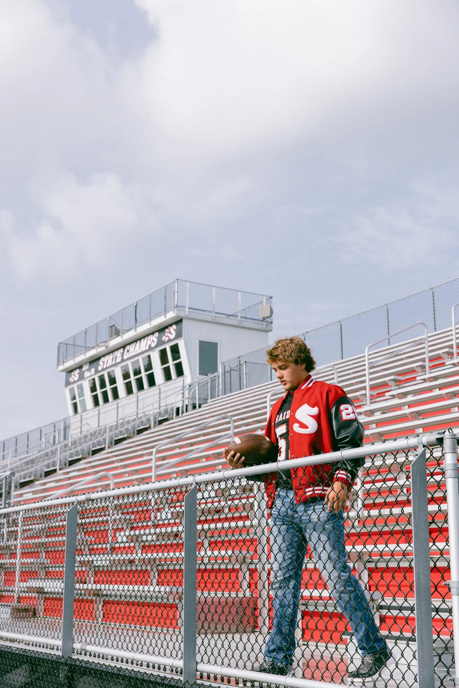 A young man with curly hair wearing a red and black varsity jacket and jeans walking down the bleachers at a football stadium, holding a football in his hands.