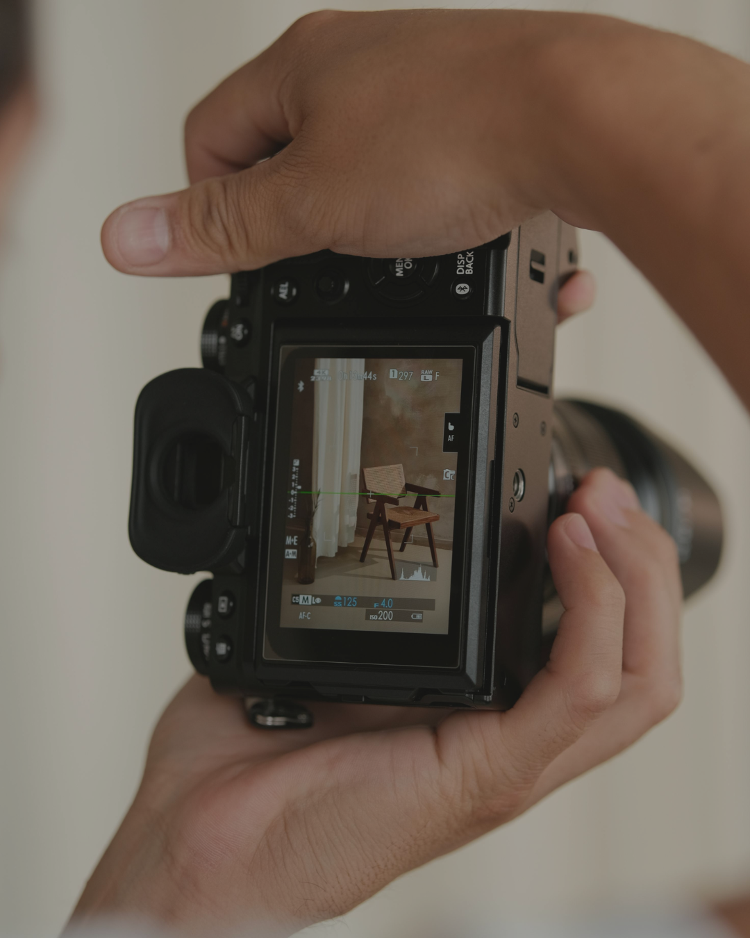 A person holding a camera, capturing an image of a wooden chair with a cushion, placed against a wall with a curtain on the left side.