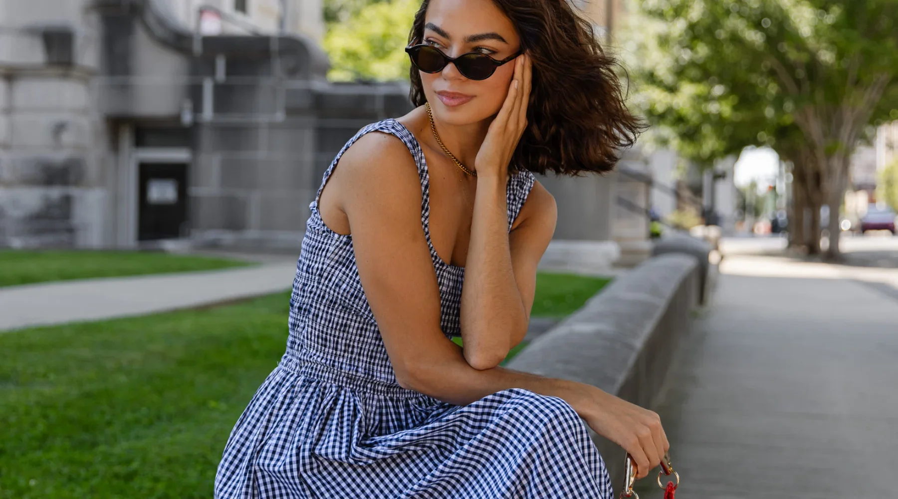 A woman with dark curly hair wearing black sunglasses, a blue and white checkered dress, and a gold necklace is sitting on a stone bench in an outdoor park, touching her face while looking to the side with a slight smile.