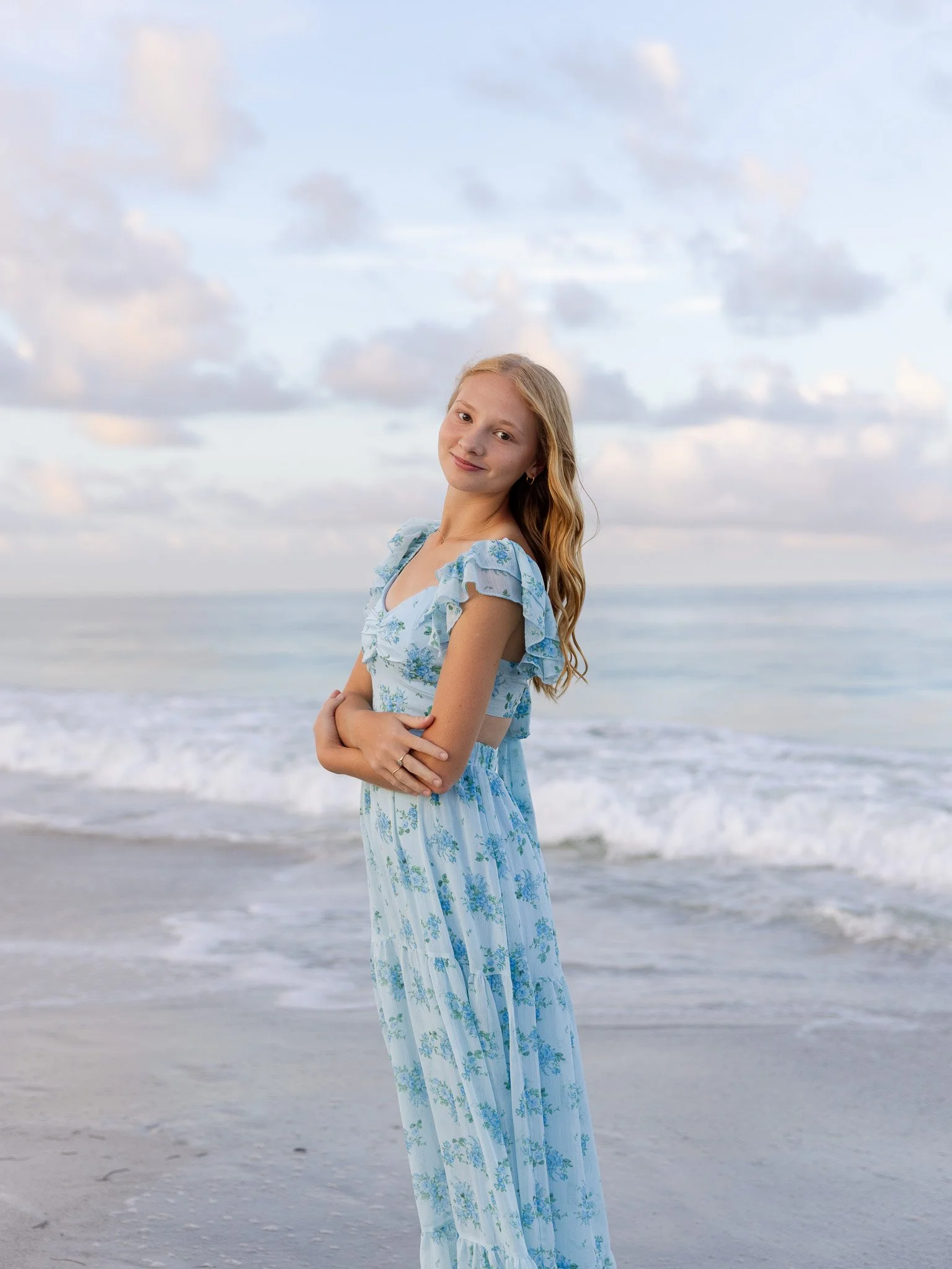 A young woman with long, wavy red hair standing on a beach in a light blue floral dress, with her arms crossed and smiling softly at the camera, ocean waves and a cloudy sky in the background.