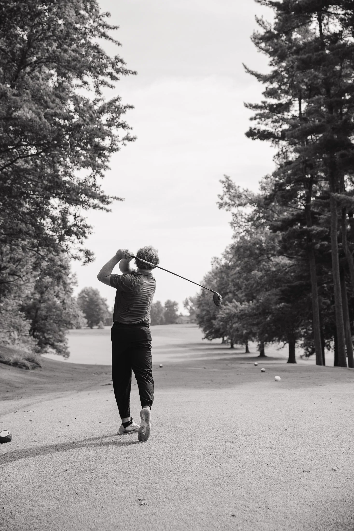 A person in a golf course preparing to hit a golf ball with a club, surrounded by trees and golf balls on the ground, in black and white.