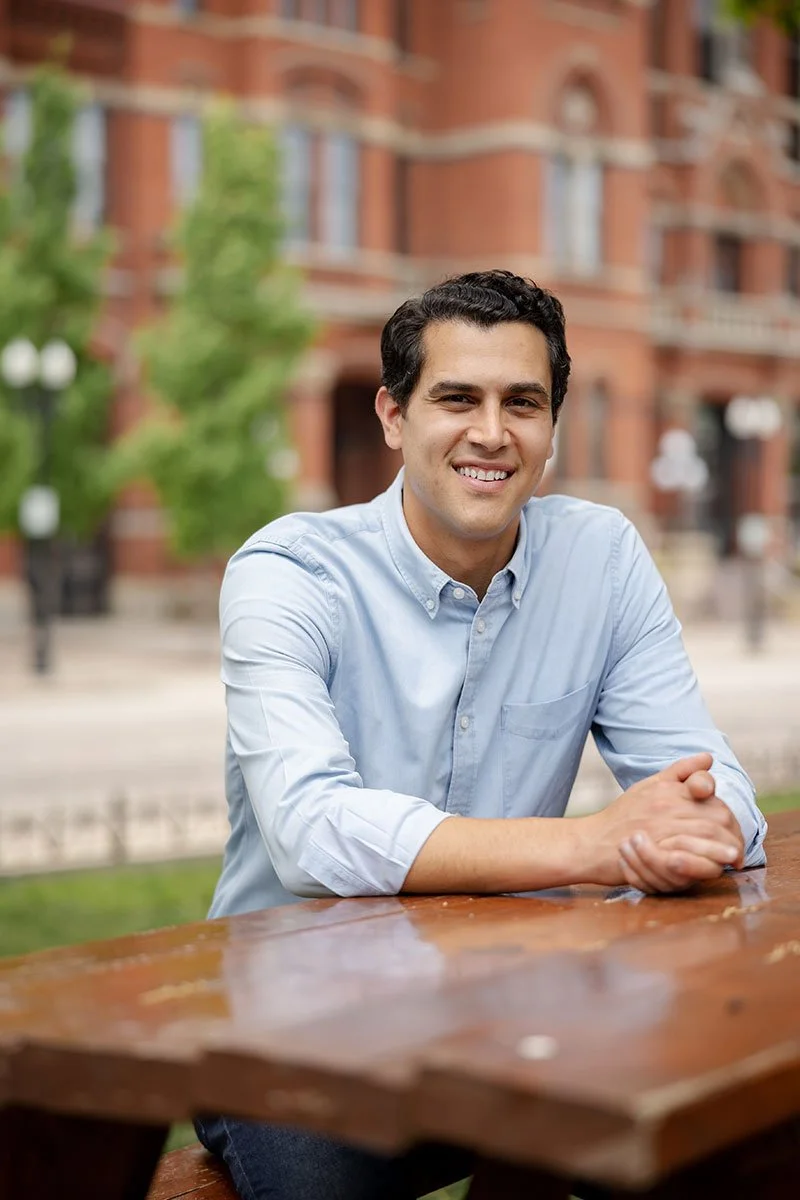 Dani Isaacsohn (a young man with dark hair, wearing a light blue button-up shirt) sits at a wooden table outdoors, smiling at the camera with a city street and brick building in the background.