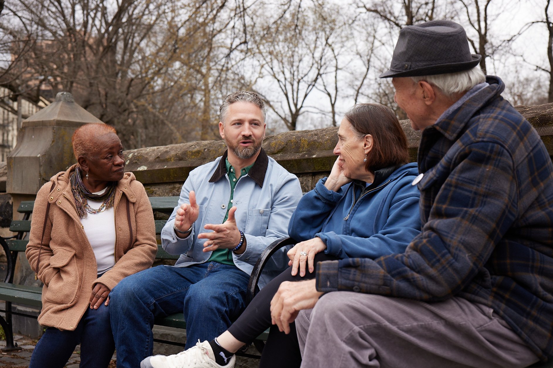 Four people sitting on a bench outdoors, engaged in a conversation, with trees and an overcast sky in the background.