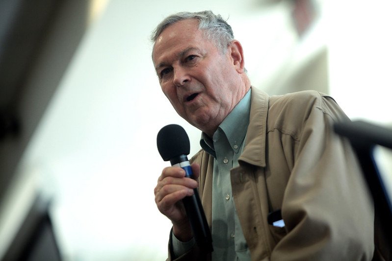 An older man holding a microphone and speaking, wearing a beige jacket and a light blue shirt, with a blurred background.