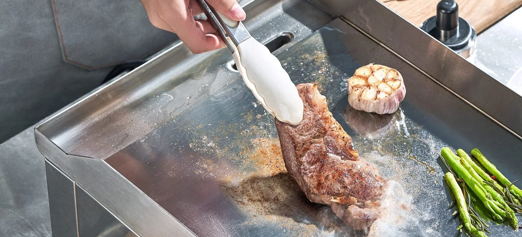 A person is placing a dollop of white sauce or cream onto a cooked piece of meat on a metal grilling surface. There is a bulb of garlic, some green vegetables, and a small bottle of oil or sauce on the grill.