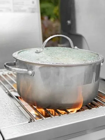 A stainless steel pot with a glass lid on a portable stove with flames underneath.