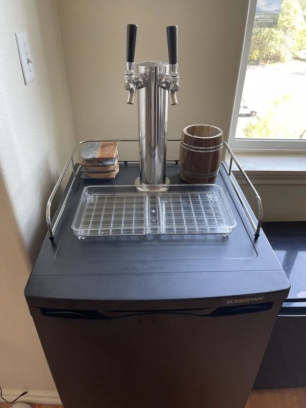 Self-serve beverage station with a dual-tap beer or soda dispenser, a clear plastic tray, a wooden cup, and a set of coasters on top of a black countertop near a window.