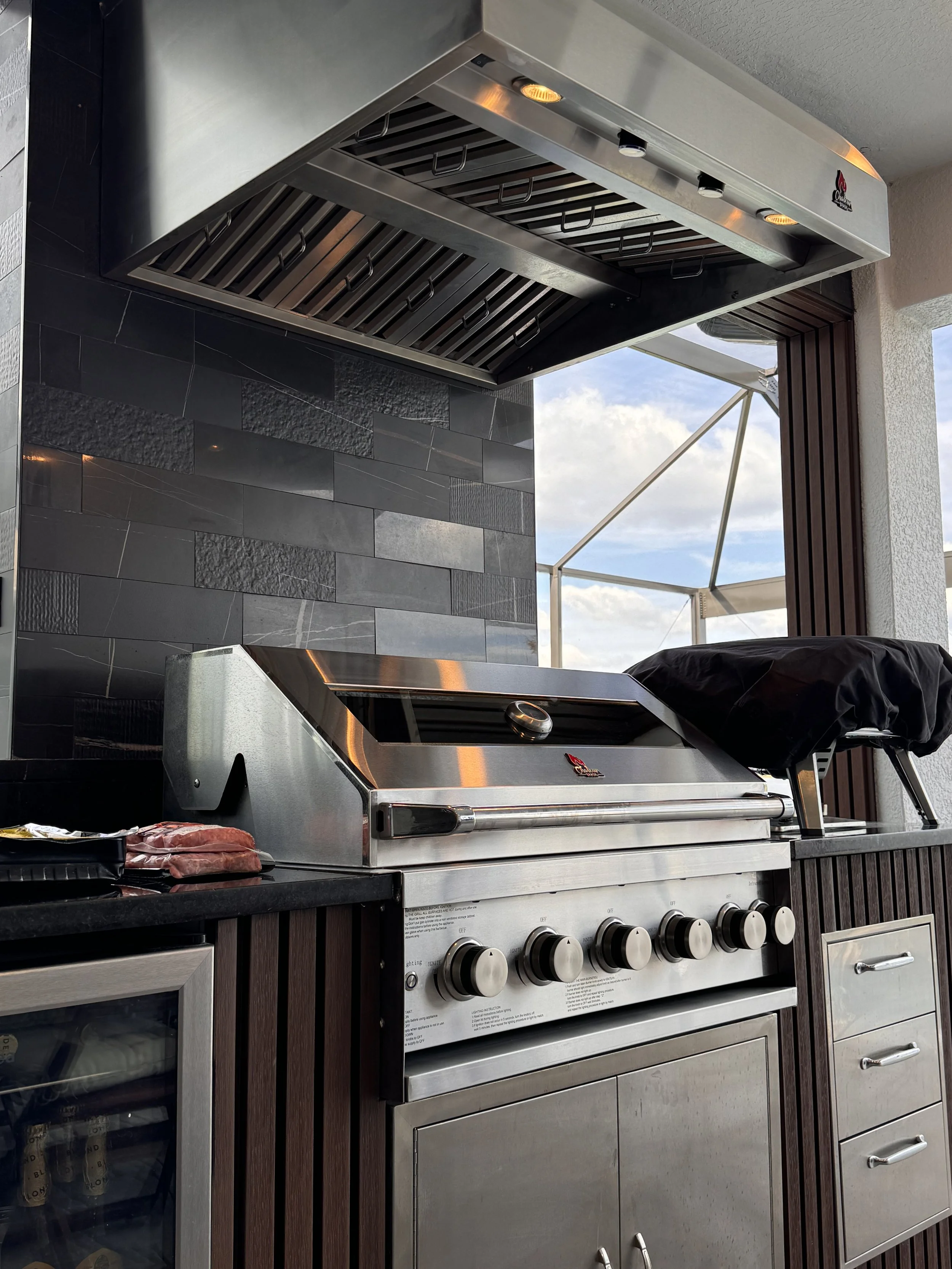 Outdoor stainless steel gas grill with six control knobs, a black cover, and a chipotle logo, situated on a wooden and stone kitchen counter, with a black tile wall and outdoor sky in the background.