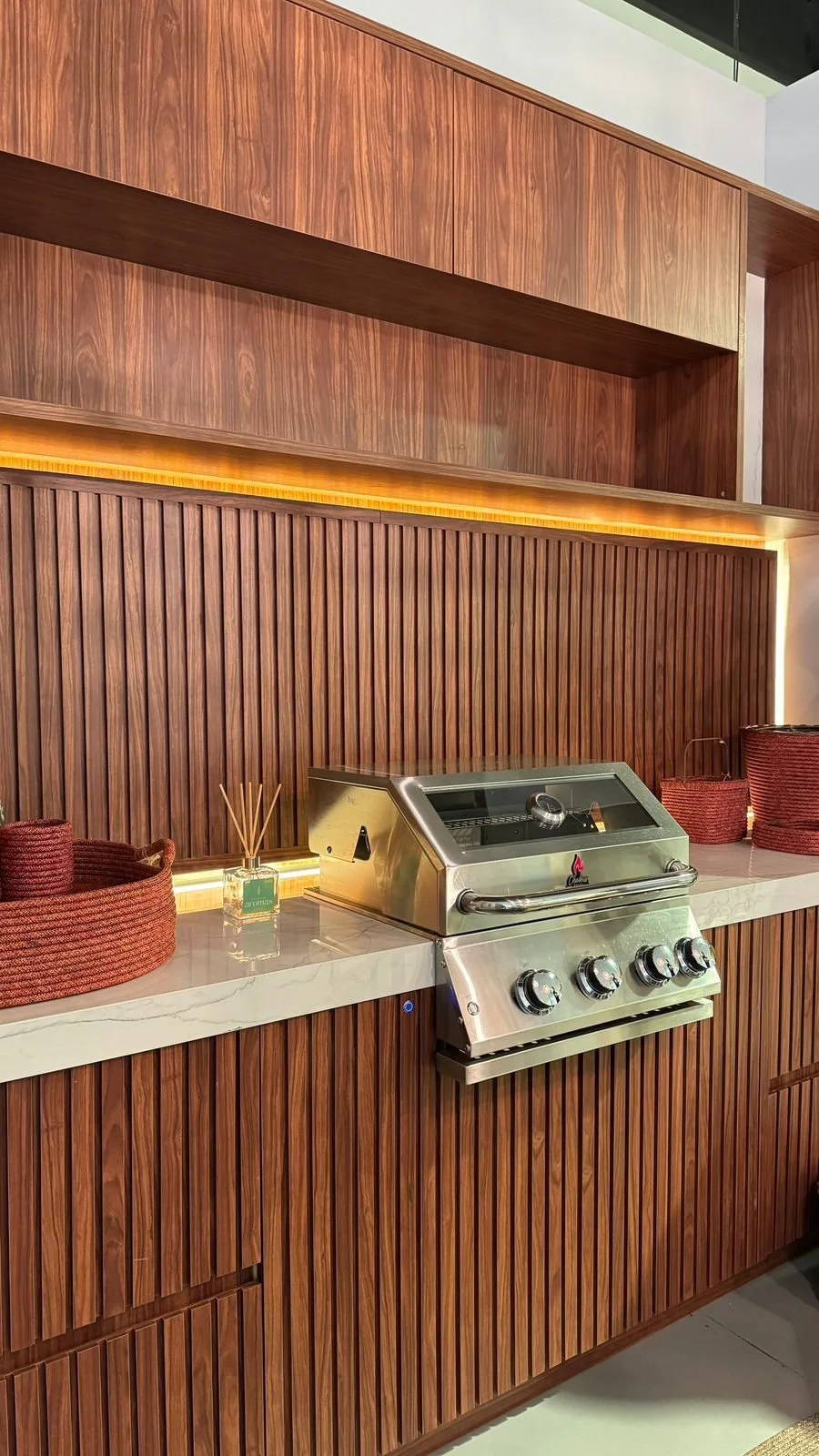 A kitchen countertop with a stainless steel grill, red woven baskets, and a diffuser with sticks, set against a wood-paneled wall with vertical slats and warm lighting.