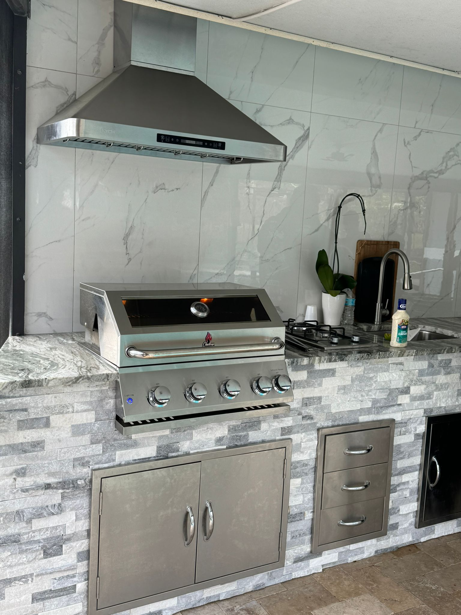 Kitchen outdoor grill with stainless steel hood, gray stone countertop, potted plant, sink with dish soap, and drawers and cabinets.