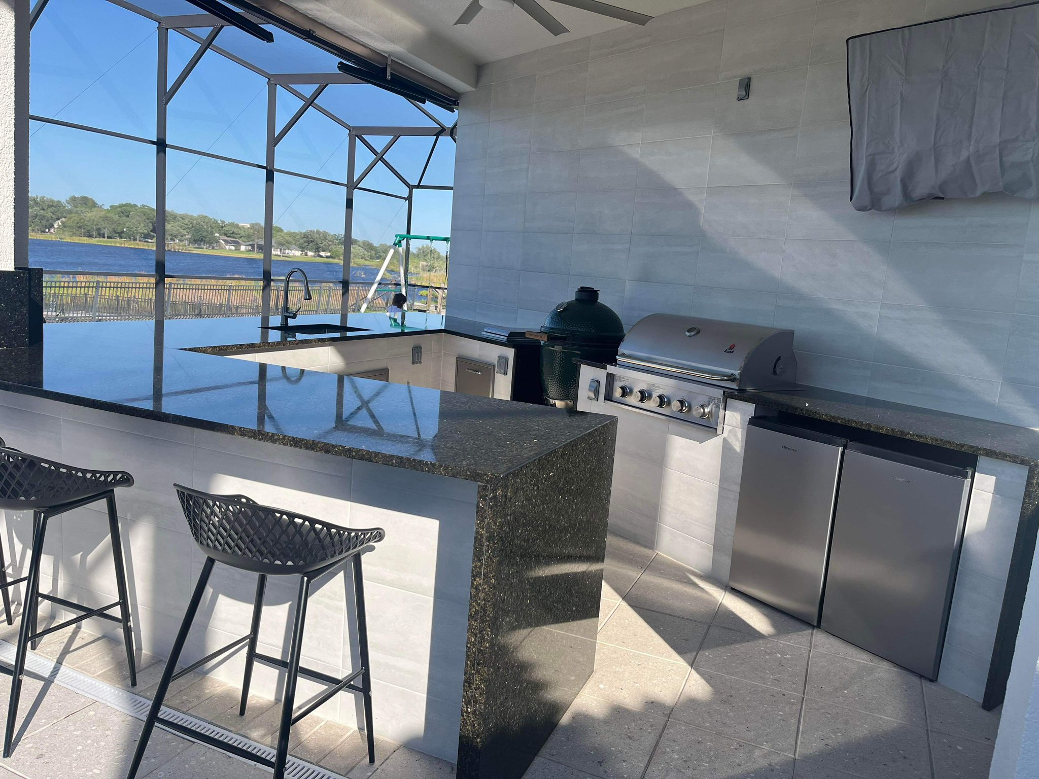 Outdoor kitchen with a granite countertop, a grill, a small fridge, and bar stools, overlooking a lake.