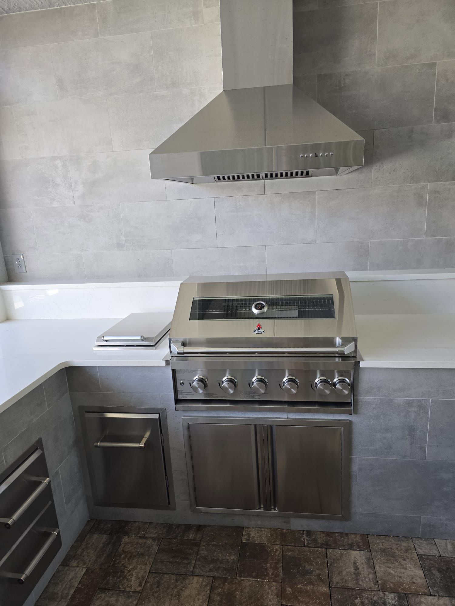 Stainless steel outdoor grill with four control knobs, a lid with a thermometer, and a grill cover. Above it, a stainless steel range hood against a gray tiled wall in a modern kitchen or patio.