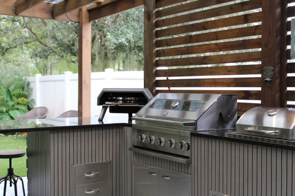 Outdoor kitchen with stainless steel grill and small side burner, wooden privacy fence, and outdoor dining area with chairs and table, surrounded by greenery.