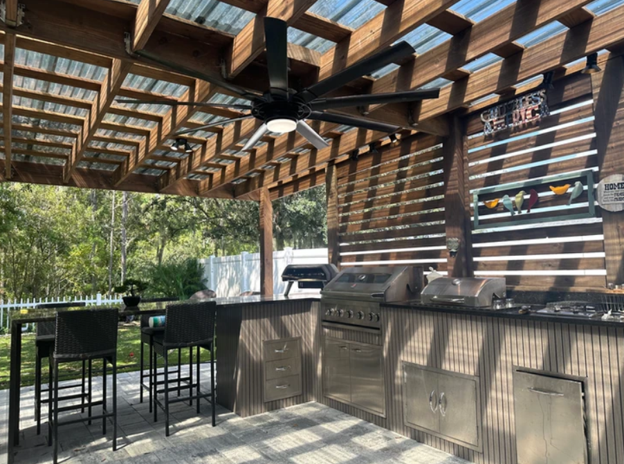 Outdoor kitchen with stainless steel grill and countertop, ceiling fan, wooden pergola, and seating area with chairs and table, surrounded by trees and a white fence.