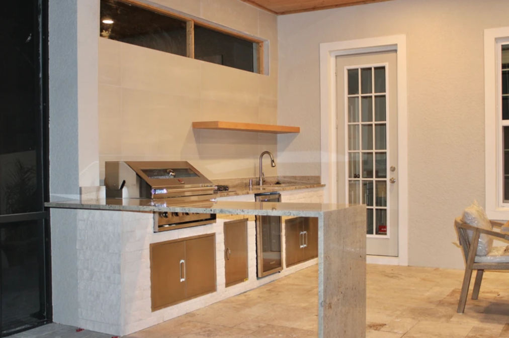 Outdoor kitchen area with a built-in grill, a sink, and a granite countertop. There is a door with glass panels and a window nearby. A chair with a plastic bag on it is visible on the right side.