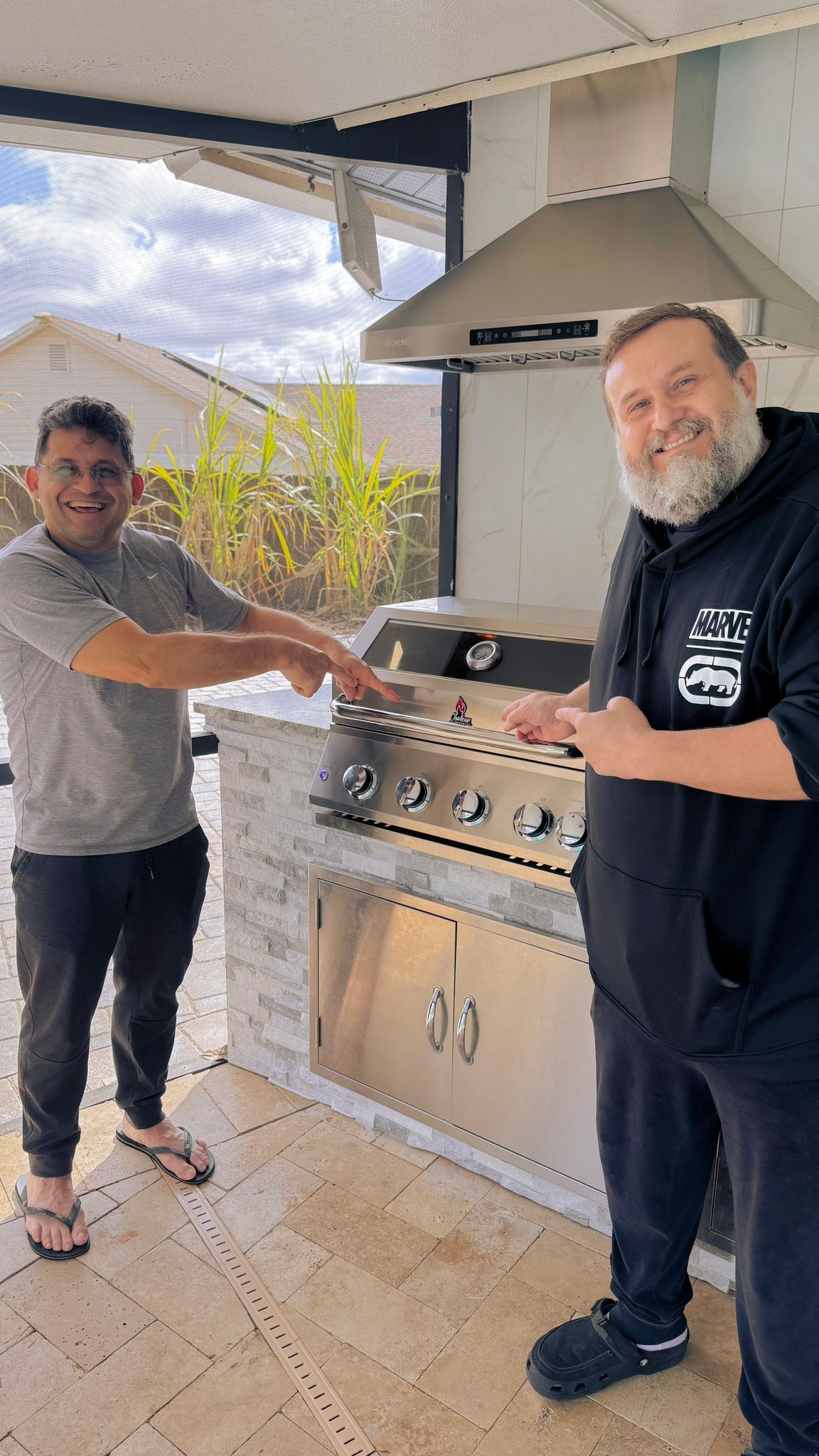 Two men standing by a stainless steel outdoor grill, smiling and pointing at it. One man has a beard and is wearing a black Marvel hoodie, the other is wearing a grey T-shirt and sandals. They are outside on a paved patio with a house and plants in the background.