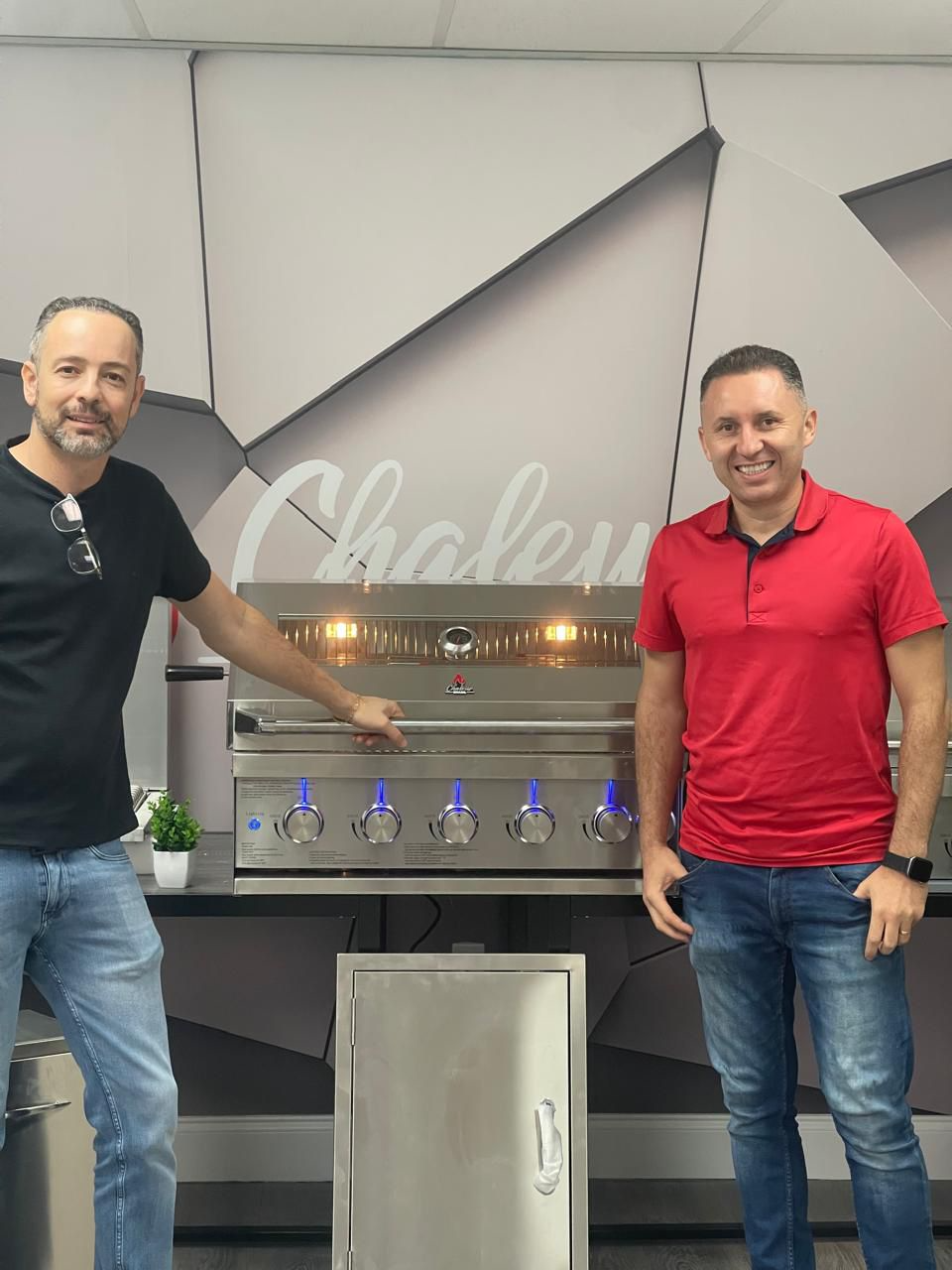 Two men standing in front of a large stainless steel grill in a modern kitchen or showroom, smiling at the camera.