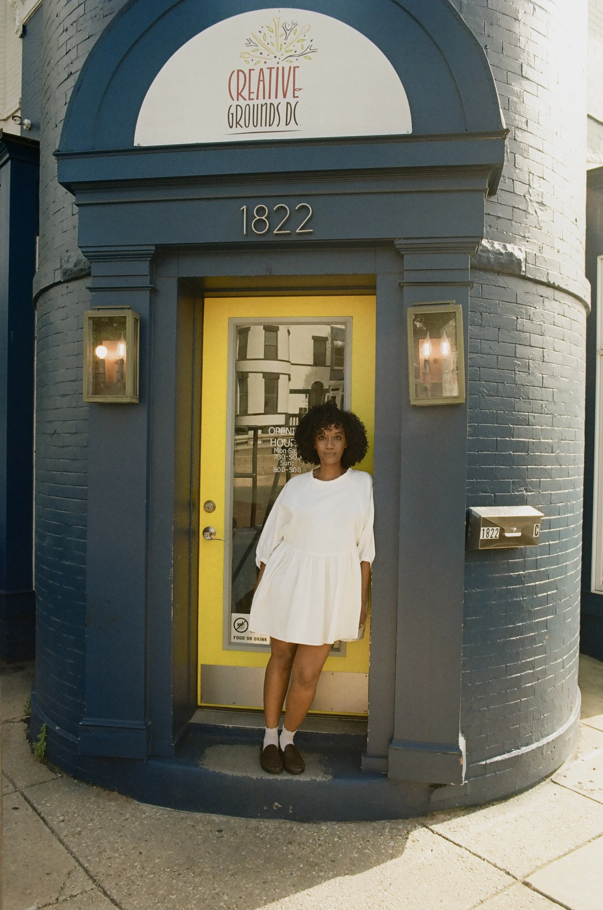 A woman in a white dress stands in front of a small blue and yellow booth at 1822. The booth has a yellow door with a window that displays hours of operation and a sign indicating food or drink. The woman has curly hair and is wearing brown shoes and socks, with her left leg slightly raised. The background shows part of a brick building and a mailbox with the number 1822.