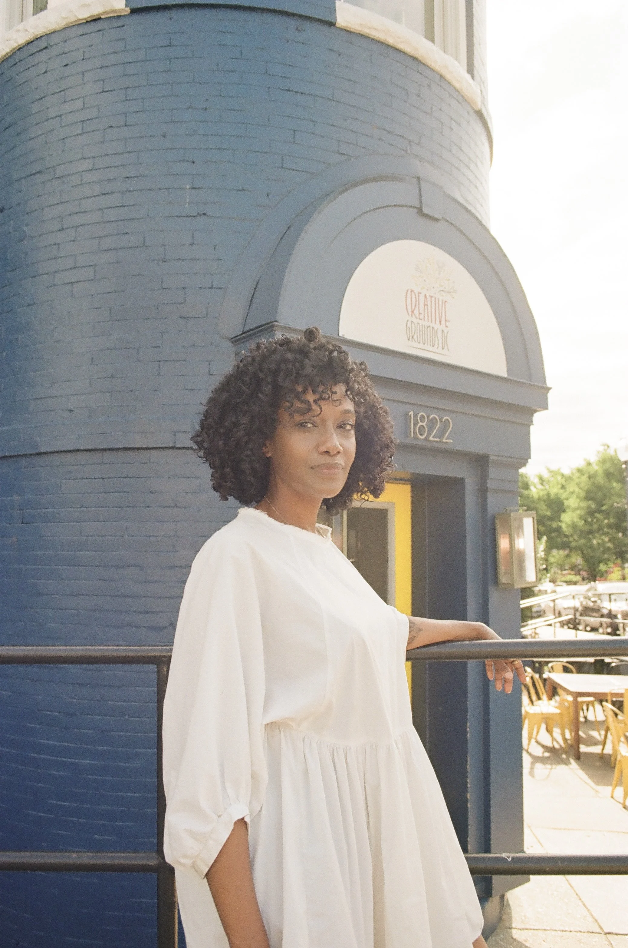 A woman with curly hair dressed in a white dress, standing outside an establishment with a blue brick corner and a sign that reads 'Creative Grounds DC' with the number 1822.