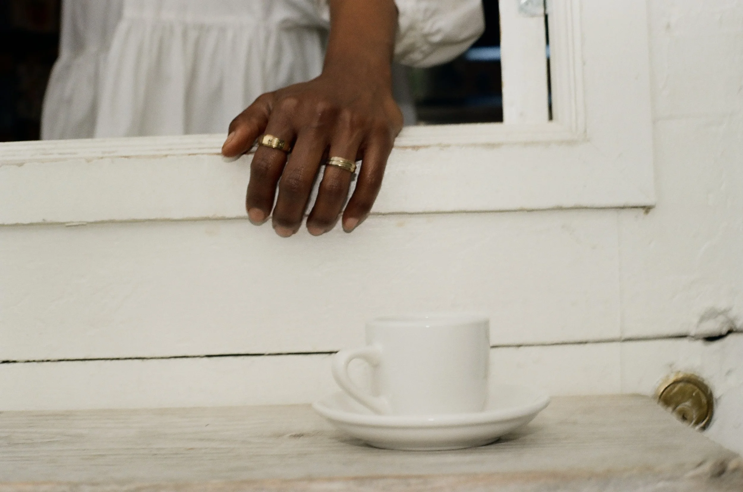 Close-up of a person's hand with rings, resting on a white window frame, with a white ceramic cup on a wooden surface below.
