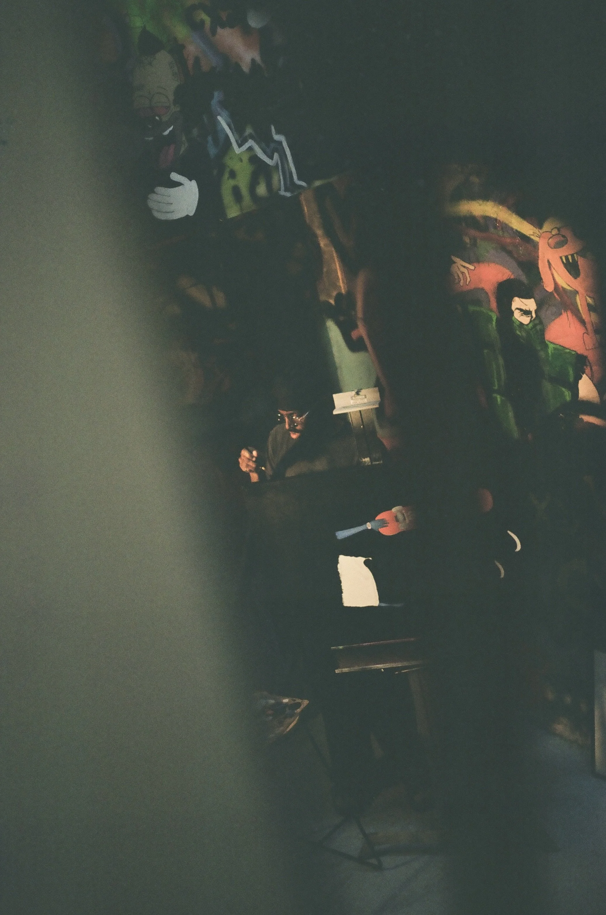 Dark room with colorful graffiti-style artwork on the wall, a person holding a drink, and some lighting equipment visible, creating an artistic and vibrant atmosphere.