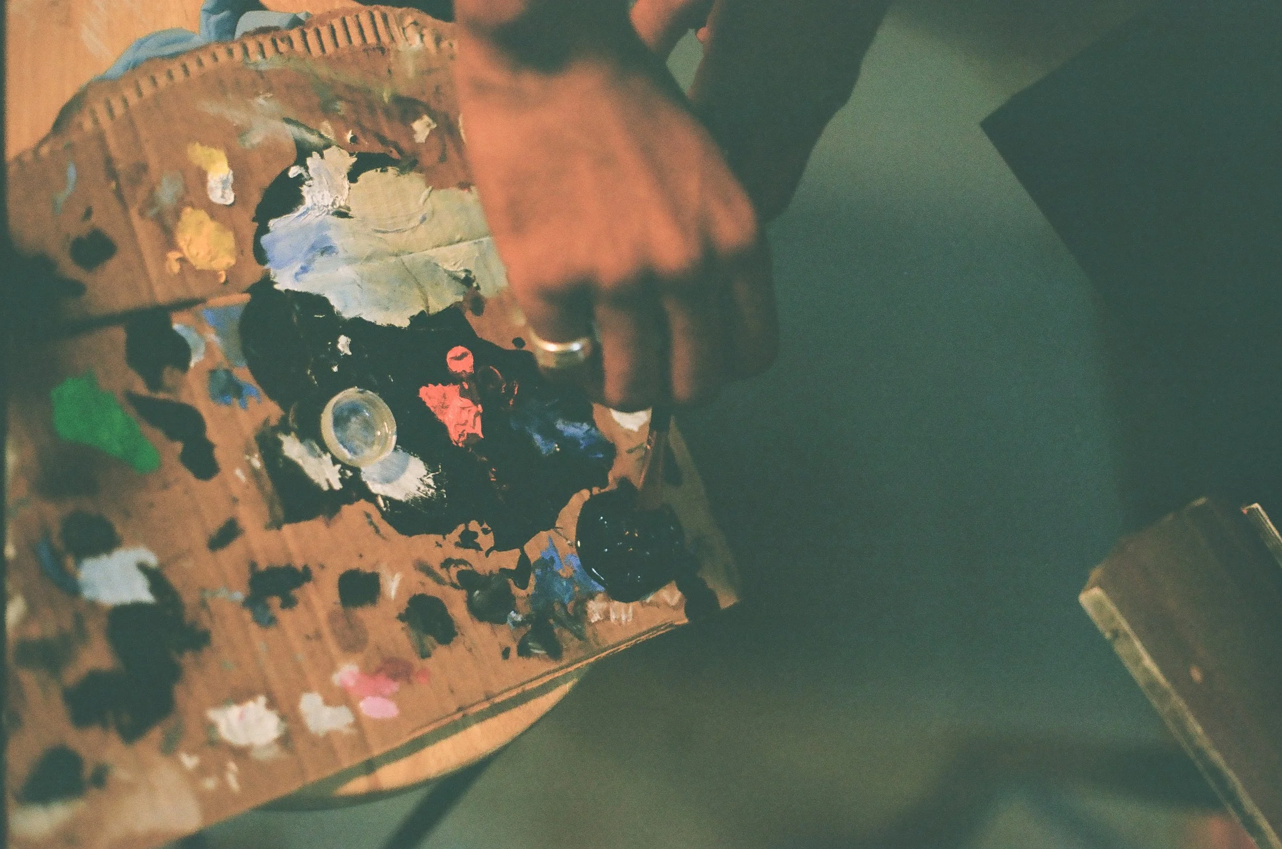 A painter's palette with various blobs of paint in colors such as black, white, red, green, blue, and yellow, resting on a wooden surface with scattered paint smudges and a person’s hand nearby.