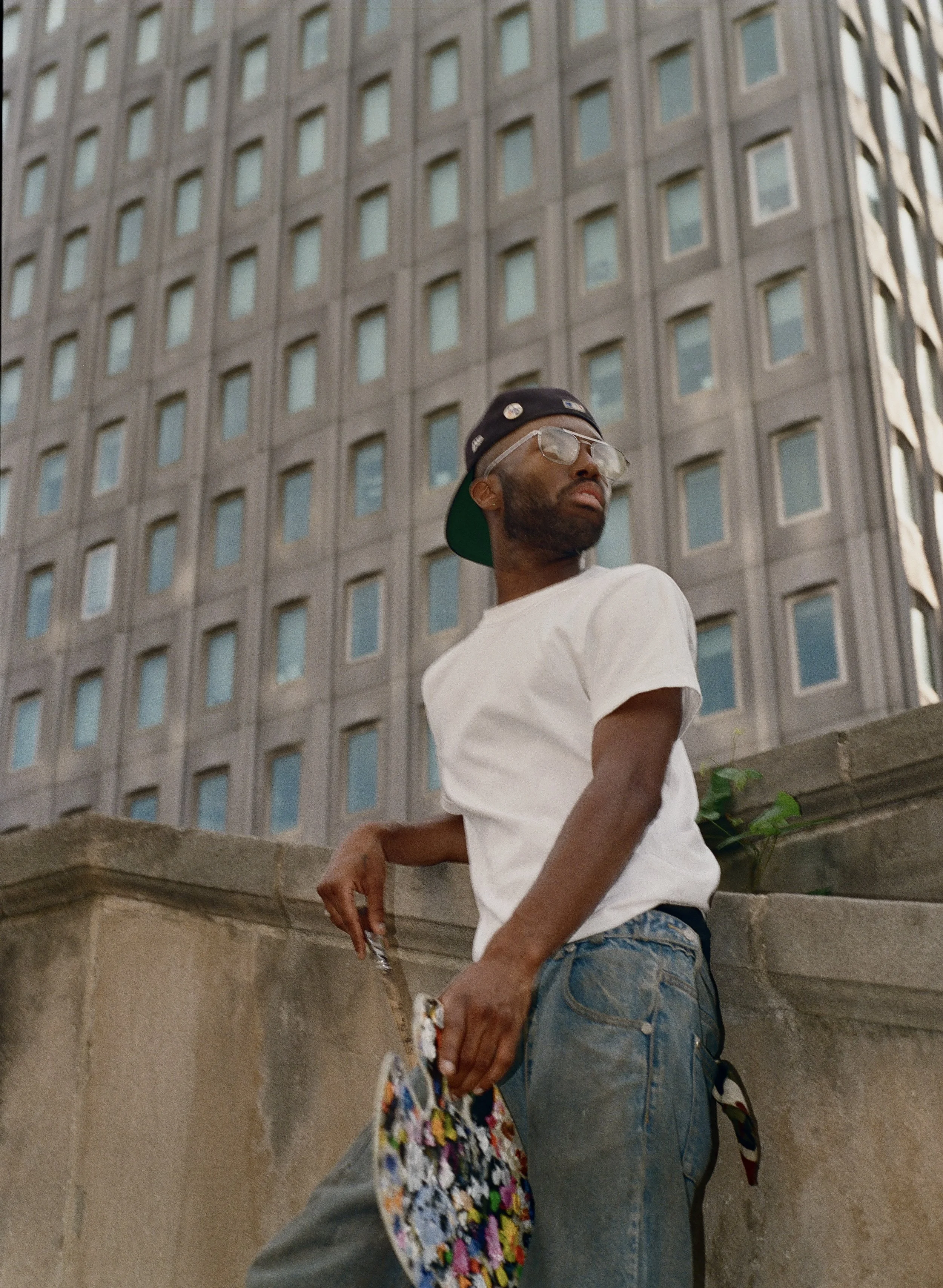 A young man wearing a black cap, sunglasses, a white t-shirt, and jeans standing outdoors in front of a tall building with many windows.