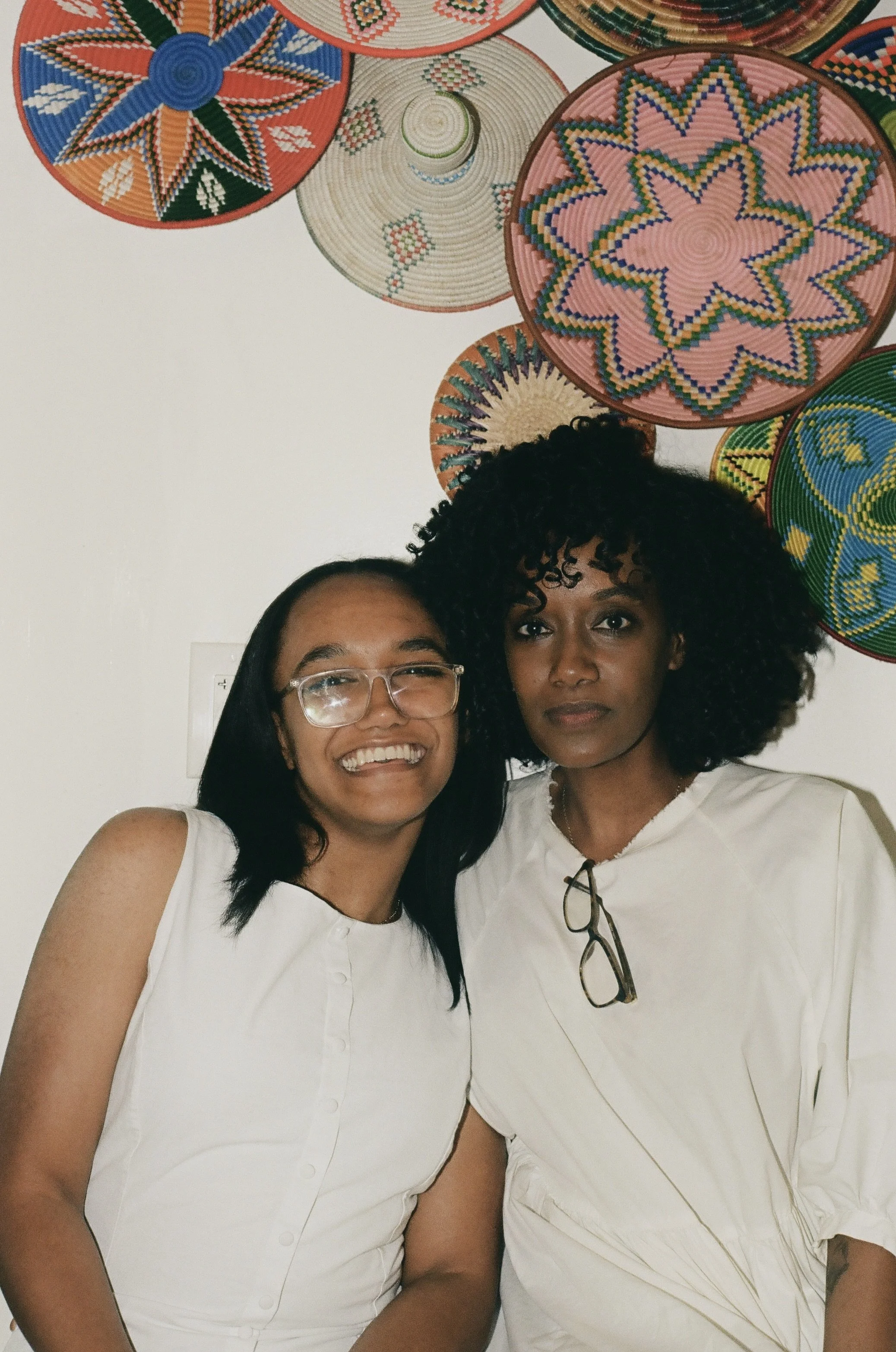 Two women standing close together, smiling, against a colorful wall decorated with circular woven baskets with intricate geometric and floral patterns.